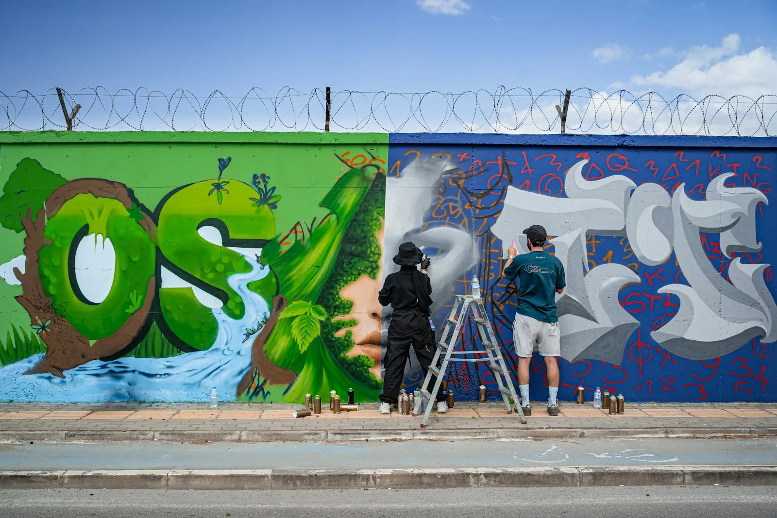 Two street artists working together on a large green and blue mural.